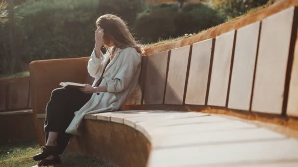 Mujer de pelo largo en gafas de sol libro de lectura en el banco en el patio. Hierba verde en primer plano. Mujer con un abrigo beige está disfrutando de un día soleado y aire fresco. Descansa en el parque. Descanso laboral. — Vídeos de Stock