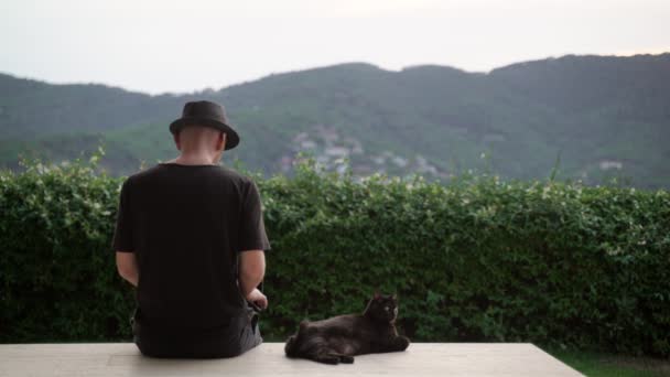 Chat noir drôle et un homme en chapeau noir se refroidissant sur la terrasse avec une belle vue. Gros chat sérieux et son ami humain. Mignon chat domestique moelleux profitant de la journée d'été. Adorable animal. Collines vertes — Video