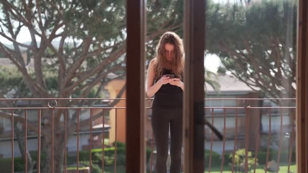 Een langharige vrouw met zwarte legging en een T-shirt staat op het balkon met een telefoon. Het meisje sms 't een bericht aan de telefoon. Mooie bomen achter haar. Communicatie. Het internet. Mobiele telefoon. — Stockvideo