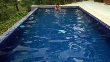 Uzun saçlı kız dinleniyor ve havuzda yüzüyor. Islak saçlı genç bir kadın yaz gününü serin mavi sularda geçirir. Yeşil ağaçların manzarası. Yaz soğuğu. Tatillerde. Tatilde. Yüzücü. 