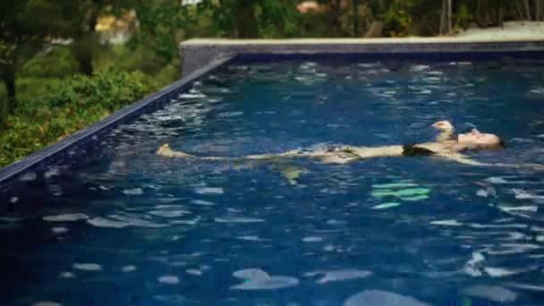 Lang haar meisje is ontspannen in het zwembad en ligt in het water. Een jonge vrouw met nat haar brengt een zomerdag door in het koele blauwe water. Uitzicht op groene bomen. Zomer chill. Vakantie. Vakantie. — Stockvideo