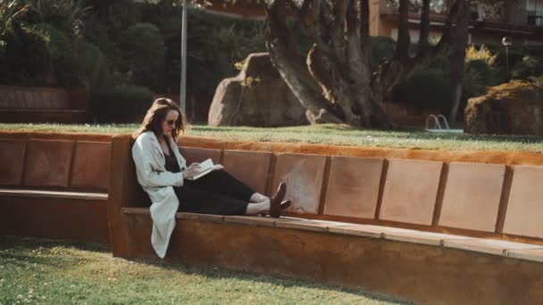 Langhaarige Frau mit Sonnenbrille liest Buch auf der Bank im Hof. Grünes Gras im Vordergrund. Frau im beigen Mantel genießt einen sonnigen Tag und frische Luft. Ruhe im Park. Arbeitspause. — Stockvideo