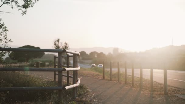 Mannen i svart joggar längs en landsväg vid solnedgången. Vacker landsbygd i gyllene ljus. Gyllene fält med trästaket och gröna kullar. En hälsosam livsstil i naturen. — Stockvideo
