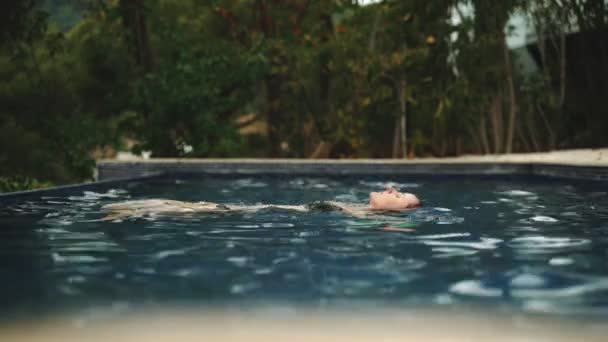 Μακριά μαλλιά κορίτσι χαλαρώνει στην πισίνα και βρίσκεται στο νερό. Μια νεαρή γυναίκα με βρεγμένα μαλλιά περνάει μια καλοκαιρινή μέρα στα δροσερά μπλε νερά. Άποψη των πράσινων δέντρων. Καλοκαίρι κρύο. Διακοπές. Διακοπές. — Αρχείο Βίντεο