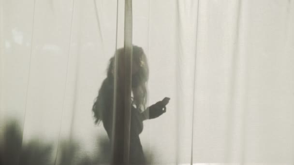 Sylwetka długowłosej dziewczyny stojącej za zasłonami smartfonem. Kontrastowy cień w jasnym słońcu. Letni nastrój. Młoda kobieta na wakacjach. Osoby wysyłające wiadomości tekstowe i dzwoniące. — Wideo stockowe