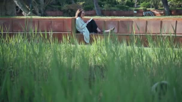 Een langharige vrouw met een zonnebril die een boek leest op de bank op de binnenplaats. Groen gras op de voorgrond. Vrouw in een beige jas geniet van een zonnige dag en frisse lucht. Rust uit in het park. Werkonderbreking. — Stockvideo