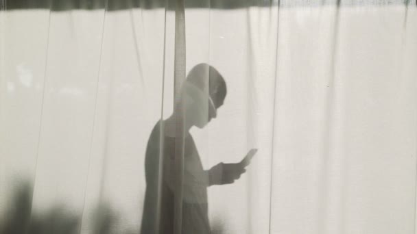Sylwetka śmiałego mężczyzny stojącego za zasłonami ze smartfonem. Kontrastowy cień w jasnym słońcu. Letni nastrój. Młody człowiek na wakacjach. Osoby wysyłające wiadomości tekstowe i dzwoniące. — Wideo stockowe