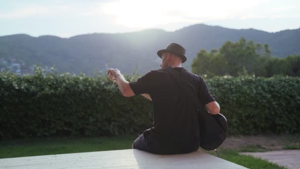 Homme en t-shirt noir et chapeau noir joue de la guitare basse. Musicien sur la terrasse avec une belle vue. Montagnes vertes et plantes en arrière-plan. Basse joueur profite de la soirée dorée. — Video