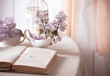 İçi leylak çiçekli vazo ve eski moda açık peri masalı kitaplarıyla dolu masa ve perdeler duvarda, yumuşak sabah ışığında romantik bahar dekorasyonu, edebiyat konsepti okuması..