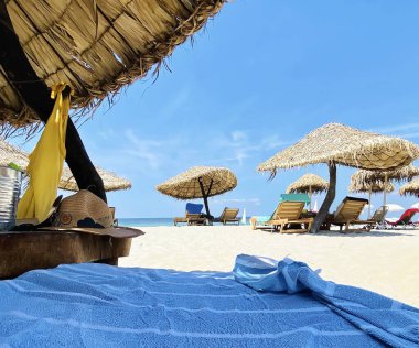 saman şemsiyeler ve ahşap sandalyeler, masa mavi battaniyesi, gökyüzü denizi, önünde sarı çiçekler, Yunanistan 'da plaj tatil beldesinde beyaz kum bulutlu sku 