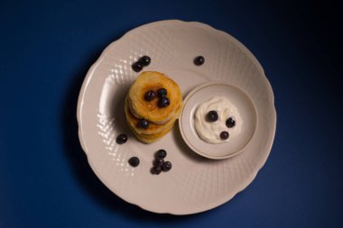 Beyaz bir tabakta peynir ve undan yapılmış küçük gözlemeler. Mavi arka planda lezzetli yemekler var.