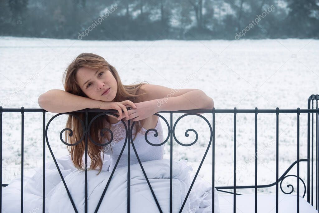young, sexy, seductive, brunette woman sitting in the cold snowy winter nature in bed, arms and pretty head laid on the iron bedstead in thought, copy space