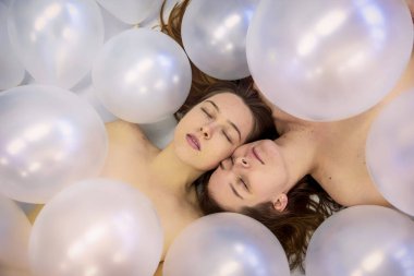 Açık gri zemin üzerinde yatarken gözleri kapalı beyaz yanardöner balonların arasında saklanan iki çekici genç kadın.