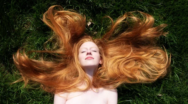 Top view to beautiful portrait of young sexy redhead woman, lying contend in the spring, summer sun, relaxing on the green meadow grass, the long red hair draped freely around the head, copy space