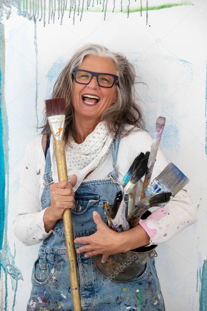 lucky portrait of an older mature woman, artist, in her fifties with grey hair and black glasses, and many big paintbrushes,