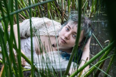 Yeşil kamışların arasında sığ sularda uzanan genç bir kadın, sakin, yoğun bakışlarla kameraya bakıyor. Hafif dantelli bir elbise giyiyor. Gizemli doğa bağlantılarını ve sessiz dayanıklılığını gösteriyor..