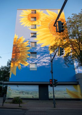 Parlak, büyük ayçiçeği duvar resmi Wuppertal 'da güneş ışığı altında yüksek binaların ön cephesini kapsıyor. Mavi arkaplan sarı yapraklarla canlı bir tezat oluştururken, bir pastoral duvar tabanı boyunca uzanır..