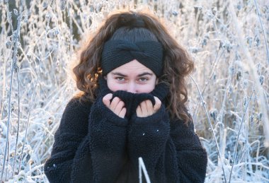 Sıcak yün ceketli genç bir kadın yumuşak sabah ışığıyla buzlu çayırda duruyor. Soğuk bitkiler ve kış sükunetiyle çevrili, neşeli bir bakışla dışarı bakıyor..