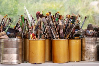 İyi kullanılmış boya fırçaları, yeşil bahçe manzarasına karşı kurumuş renklerle kaplı bir stüdyo penceresinin üzerinde sarı ve gümüş kutularda duruyor ve yaratıcı bir atmosfer yaratıyor..