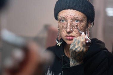 Yüzüne odaklanmış yoğun bir ifadeyle kına çilleri sürerken siyah bir bere takan ve piercing takan genç bir kadının yakın portresi.