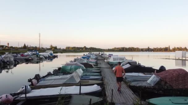 Een man loopt op de steiger tussen de boten, in een rivierbaai. Triatleet Hardlopen, Sprinten en uithoudingsvermogen training. Hij jogt door het hele land. Training en buitensporten. Langzame beweging — Stockvideo