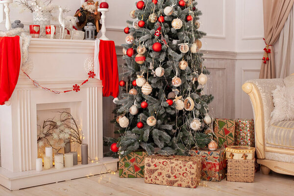 Decorated Christmas tree with gifts, fireplace and candles. Christmas tree decorated with toys and lights glowing garlands in living room. Xmas decoration concept. Cozy xmas interior room with sofa