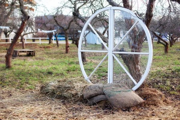 Exterior decor of wedding ceremony in a rustic style. Large wooden wheel with bags and hay next  trees in the garden. Beautiful garden decor outdoors. Decorative White wheel in apple orchard.