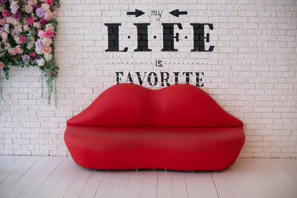 Red sofa lips shape on white brick wall background. Minimalism concept. Sofa lips in white room and text on wall. Lips shape sofa in floral design. Living room interior with modern red sofa