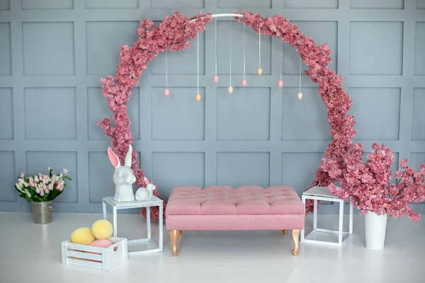 Spring home interior with pink sofa, blossom large sakura wreath on wall. Modern interior decor home. Children room in Easter style. Easter decoration of living room with rabbits and box with eggs.