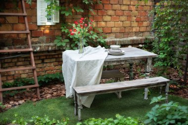 Teras evinin önünde tuğla duvar ve bitkilerle, masa ve ahşap bankla. Bahçe, veranda, ahşap bahçe mobilyası ve barbekü. Arka bahçede yemek masası. Avlu ya da balkonda rahat bir yer. 