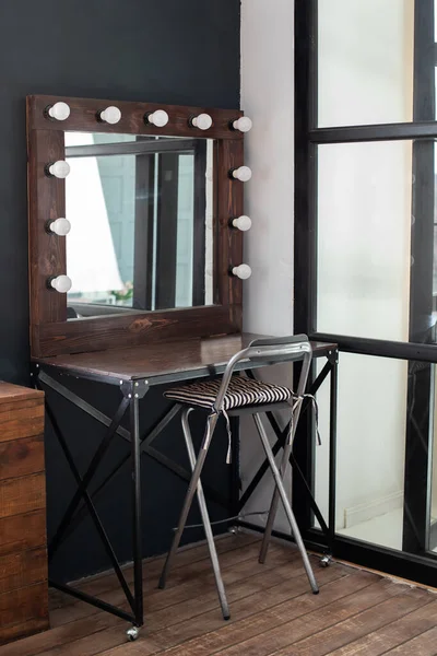 Table with mirror near dark wall. Dressing room with makeup mirror and table. Hairdresser place for hairdresser and make-up artist - seats and illuminated mirror. Modern loft interior makeup room.