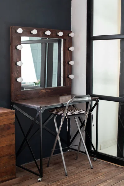 Table with mirror near dark wall. Dressing room with makeup mirror and table. Hairdresser place for hairdresser and make-up artist - seats and illuminated mirror. Modern loft interior makeup room.