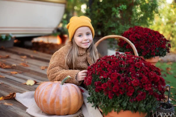 在秋天的花园里用黄色的南瓜和花拍打孩子 快乐的小女孩坐在门廊上 手里拿着菊花 盆栽和南瓜 万圣节或感恩节的家居装饰 — 图库照片