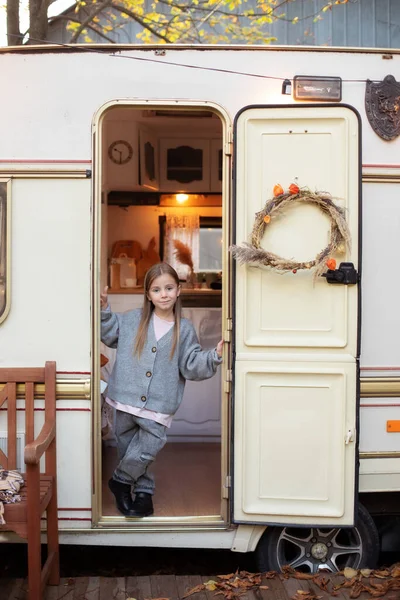 Smiling little girl in casual clothes standing on porch RV house in garden. Cute young girl stand near trailer door. Child in cozy campsite fall backyard. Concept camping, outdoor, nature, adventure