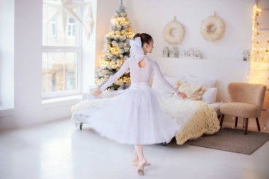 Genç bir balerin, şenlikli bir yatak odasında arka planda dans eder evde etek ve parlak çelenklerle Noel ağacının yanında sivri ayakkabılar giyer ve rahat bir tatil atmosferi yaratır. 