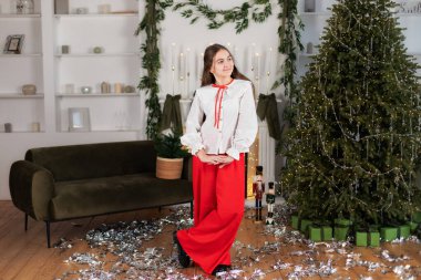 Noel 'de bir salonda süslenmiş Noel ağacı çelengi ışığı ve hediyelerle duran zarif görünümlü genç bir kız. Kırmızı pantolonlu genç bir kız ve yeni yılda süslü bir evde beyaz bir gömlek.. 