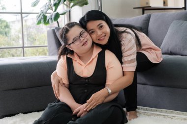 Asyalı bir anne otizmli genç bir kızı kucaklıyor gülümsüyor ve evde yerde kameraya bakıyor. Otistik genç öğrenciler öğretmenlerle öğreniyor..