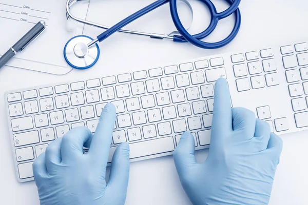 Doctor Hands in gloves typing on computer keyboard on white desk. Telemedicine or medical technology concept background. Top view