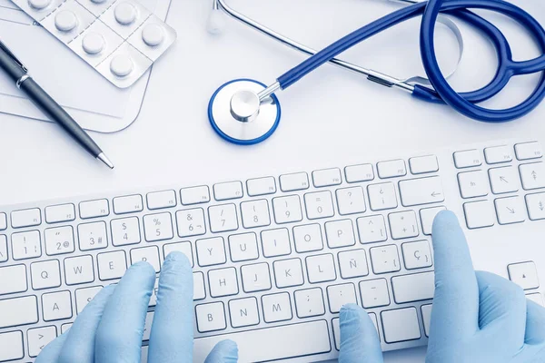 Doctor Hands in gloves typing on computer keyboard on white desk. Telemedicine or medical technology concept background. Top view