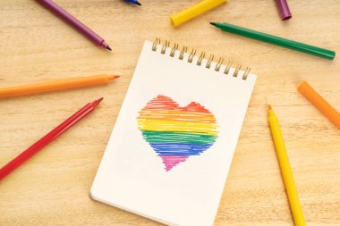 LGBT gökkuşağı bayraklı defter. Kalp şeklinde çizim ve tahta masa üzerine işaret kalemleri.