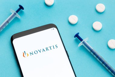 Galiçya, İspanya; 8 Haziran 2020: Ekranda Novartis logosu, mavi arka planda haplar ve şırıngayı gösteren akıllı telefon