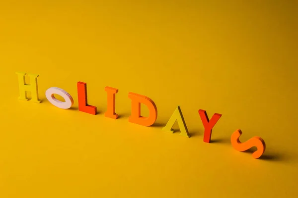 the inscription holidays from colorful bright wooden letters lie on a bright yellow background top view copy space.
