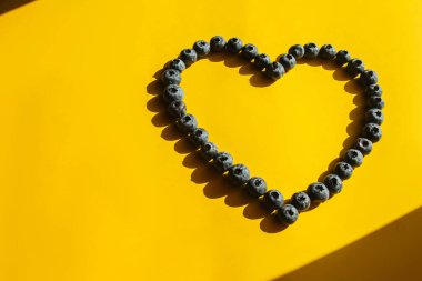 Sağlıklı yiyecekler, çiğ yiyecekler. Sarı arka planda kalp şeklinde yaban mersini. Sert gölgeler. Aşk sembolü.