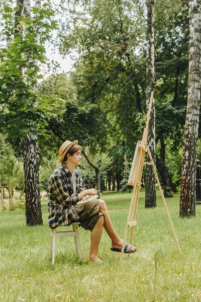 a young guy in a straw hat sits in the park in front of an easel and paints a picture with oil paints. Artistic work in nature. Plein air.