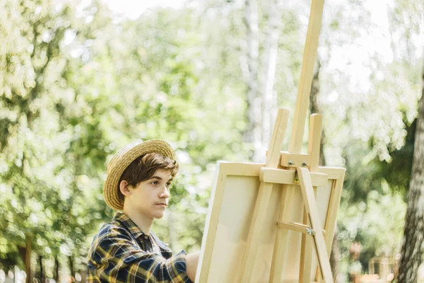 a young guy in a straw hat sits in the park in front of an easel and paints a picture with oil paints. Artistic work in nature. Plein air.