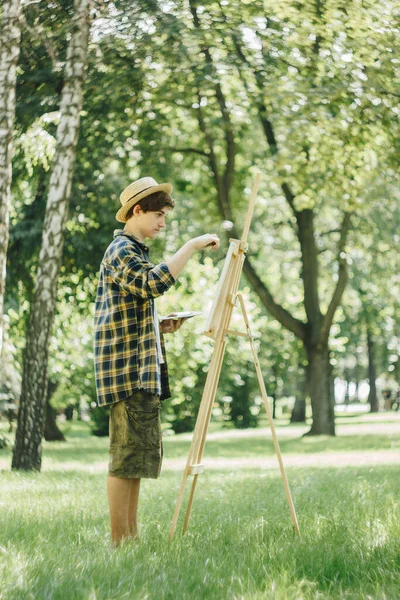 a young guy in a straw hat sits in the park in front of an easel and paints a picture with oil paints. Artistic work in nature. Plein air.