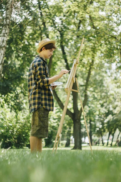 Young Guy Straw Hat Sits Park Front Easel Paints Picture — Stock Photo, Image