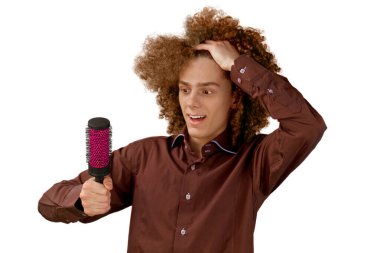 Beyaz arka planda kahverengi gömlekli, uzun saçlı, kıvırcık saçlı bir adam metal bir tarak kullanıyor. Bir kuaförde saç tıraşından önce duygular.