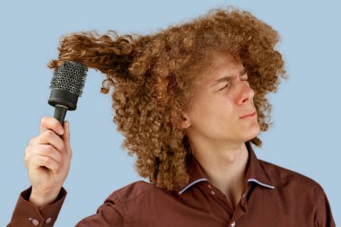 Mavi arka planda kahverengi gömlekli, uzun saçlı, kıvırcık saçlı bir adam ahşap tarak kullanıyor. Bir kuaförde saç tıraşından önce duygular. Taramaktan dolayı acı.