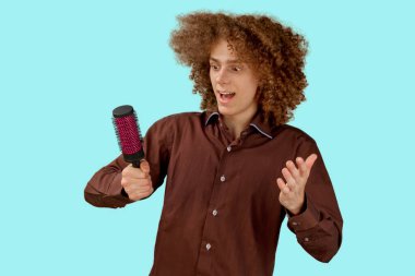 Mavi arka planda kahverengi gömlekli, uzun saçlı, kıvırcık saçlı bir adam ahşap tarak kullanıyor. Bir kuaförde saç tıraşından önce duygular.